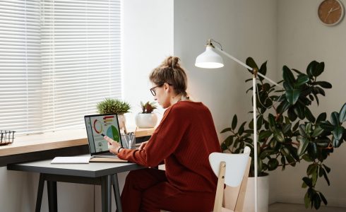 How to Buy a Home When You're Self-Employed. Rear view of woman sitting at the table with laptop and working with financial graphs online at office